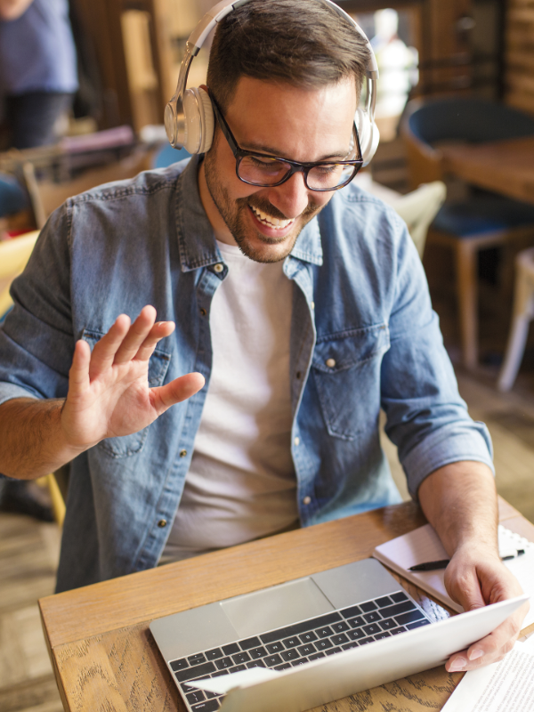 Mann mit Kopfhörern sitzt am Laptop und hebt die Hand und lacht